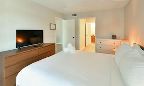 Habitación de hotel con una cama, un televisor sobre un aparador de madera, un tocador blanco y una lámpara.