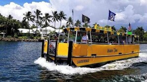 Un bote amarillo llamado 'Water Taxi' con la leyenda 'LAUDERDALE' en el costado, navegando sobre el agua con palmeras y un edificio al fondo.