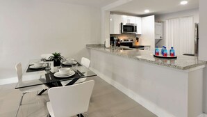 Una cocina moderna con microondas, estufa y refrigerador. Hay un comedor con mesa de vidrio y sillas blancas.