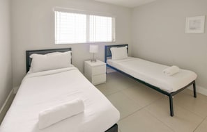 Quarto com duas camas de solteiro, mesa de cabeceira com abajur e janela com persianas.