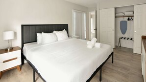 A neatly made bed with white linens, a bedside table with a lamp, and a wardrobe with hanging clothes.