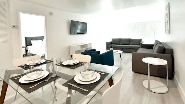 A modern living room with a dining area, a grey sofa, a flat-screen TV, and a glass dining table set for two. - ✨One Bedroom Stunning River Views at Fort Lauderdale (Fort Lauderdale)
