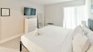 Ein Hotelzimmer mit einem Bett, einem an der Wand befestigten Fernseher, einer Kommode und einem Fenster mit Jalousien.