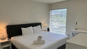 A modern hotel room with a large bed, a bedside table with a lamp, a small bedside table with a plant, and a view of the outside through a window. - ✨ New Designer One Bedroom Apartment at Fort Lauderdale (Fort Lauderdale)