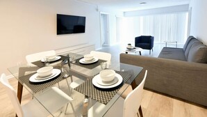 A modern living room with a glass dining table set for four, a sofa, a TV mounted on the wall, and a window with blinds. - ✨ New Designer One Bedroom Apartment at Fort Lauderdale (Fort Lauderdale)