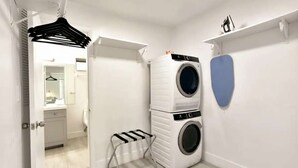 A compact laundry room with a stacked washer and dryer, a small folding chair, and a blue towel rack. - ✨ New Designer One Bedroom Apartment at Fort Lauderdale (Fort Lauderdale)