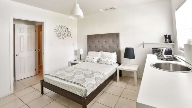 Une chambre moderne comprenant un lit, des tables de chevet, une commode, une lampe, une œuvre d’art murale et un coin cuisine avec un évier.