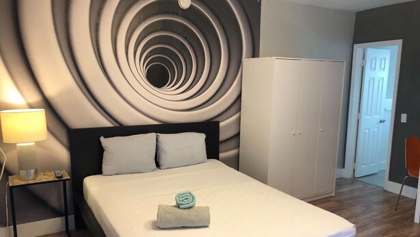 A neatly made bed with a folded towel, a bedside lamp, and a wall with a circular pattern.