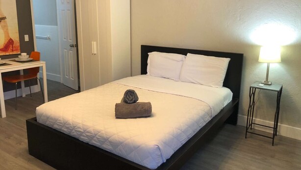 A neatly made bed with white bedding and pillows, a bedside lamp, a small table with a chair, and a wardrobe. - ✨Awesome Designer Krymwood Flat (Miami)