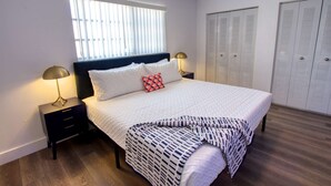 Een netjes ingerichte slaapkamer met een bed, bedlampen, een raam met zonwering en een aantal kasten.