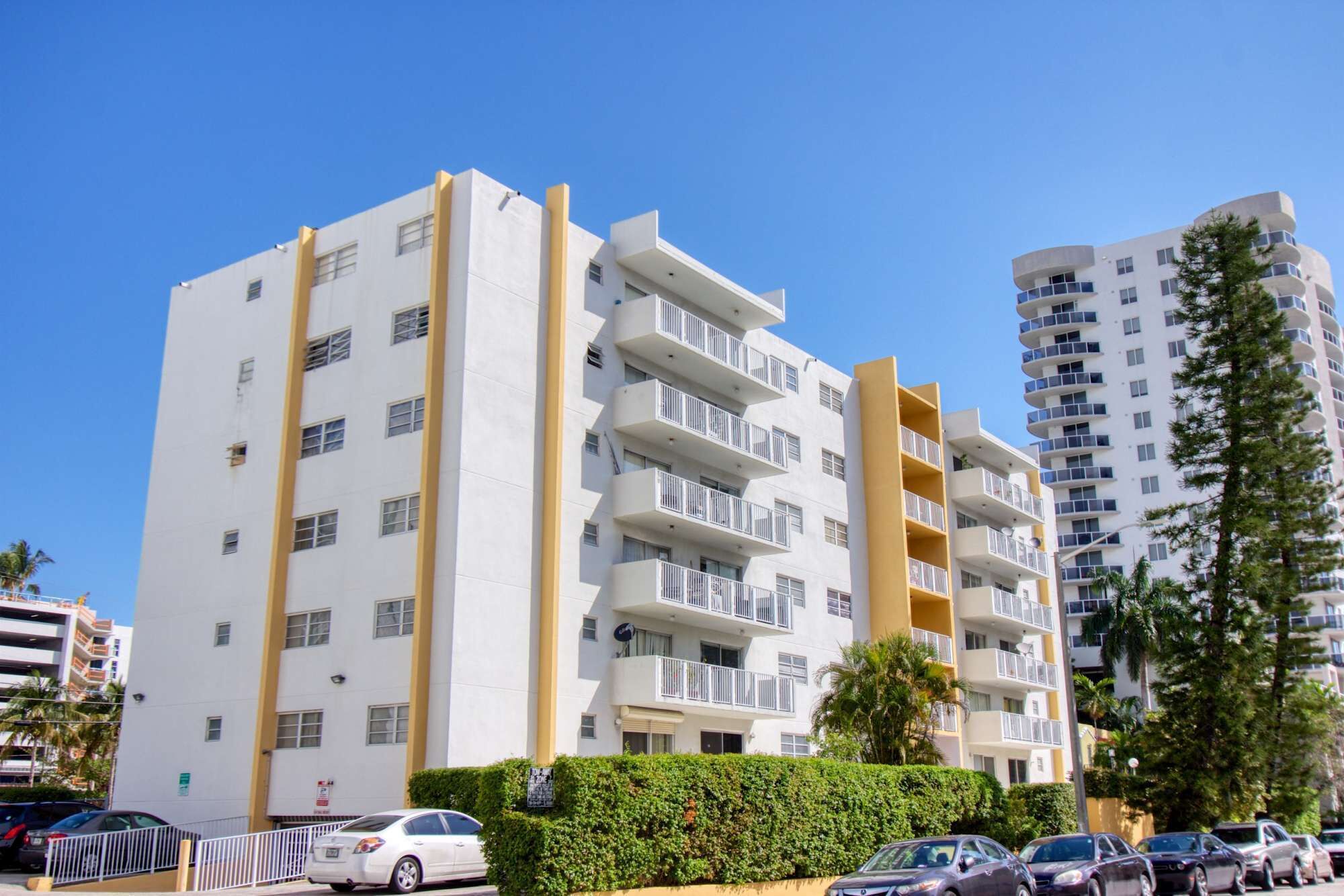 A multi-story residential building with balconies, a parking area, and a neighboring taller building.