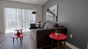 Una sala de estar moderna con un sofá gris, dos mesas rojas, una lámpara de pie y un cuadro de un paisaje urbano en la pared.