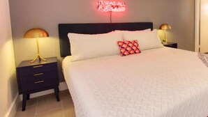 A neatly made bed with a white comforter and pillows, two bedside tables with lamps, and a neon sign on the wall. - Amazing Renovated One Bedroom w/Free Parking and Pool with Balcony (Miami)