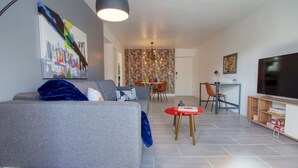 Un salon moderne avec un canapé gris, une table d’appoint rouge, un téléviseur et un coin repas agrémenté d’un papier peint à motifs.