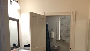 A small, compact bathroom with a sink, mirror, and towel rack. A door leads to a shower area with a curtain. - Marie Celeste Creole Country Cottage (Destrehan)