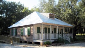 A small, single-story building with a metal roof and green shutters, surrounded by trees. - Marie Celeste Creole Country Cottage (Destrehan)