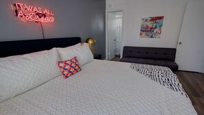 Een slaapkamer met een groot bed, een neonbord, een grijze bank en een kleurrijk schilderij aan de muur.