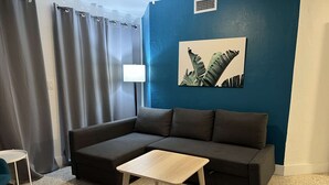 Una sala de estar con un sofá seccional gris oscuro, una pequeña mesa de madera, un sillón gris y una pared azul con una obra de arte con estampado de hojas.