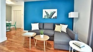 Een woonkamer met een blauwe accentmuur, een grijze bank met decoratieve kussens, twee houten bijzettafels en een kamerplant.