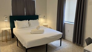 A neatly made bed with white linens and a folded towel, a dark green headboard, two bedside tables with lamps, a window with grey curtains, and a small round table. - ✨New Two Bedroom plus Free Parking (Miami)