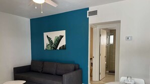 A modern living room with a dark grey sofa, a small round table, and a dining table set with white plates and glasses. A large painting of foliage hangs on the blue wall. - ✨New Two Bedroom plus Free Parking (Miami)