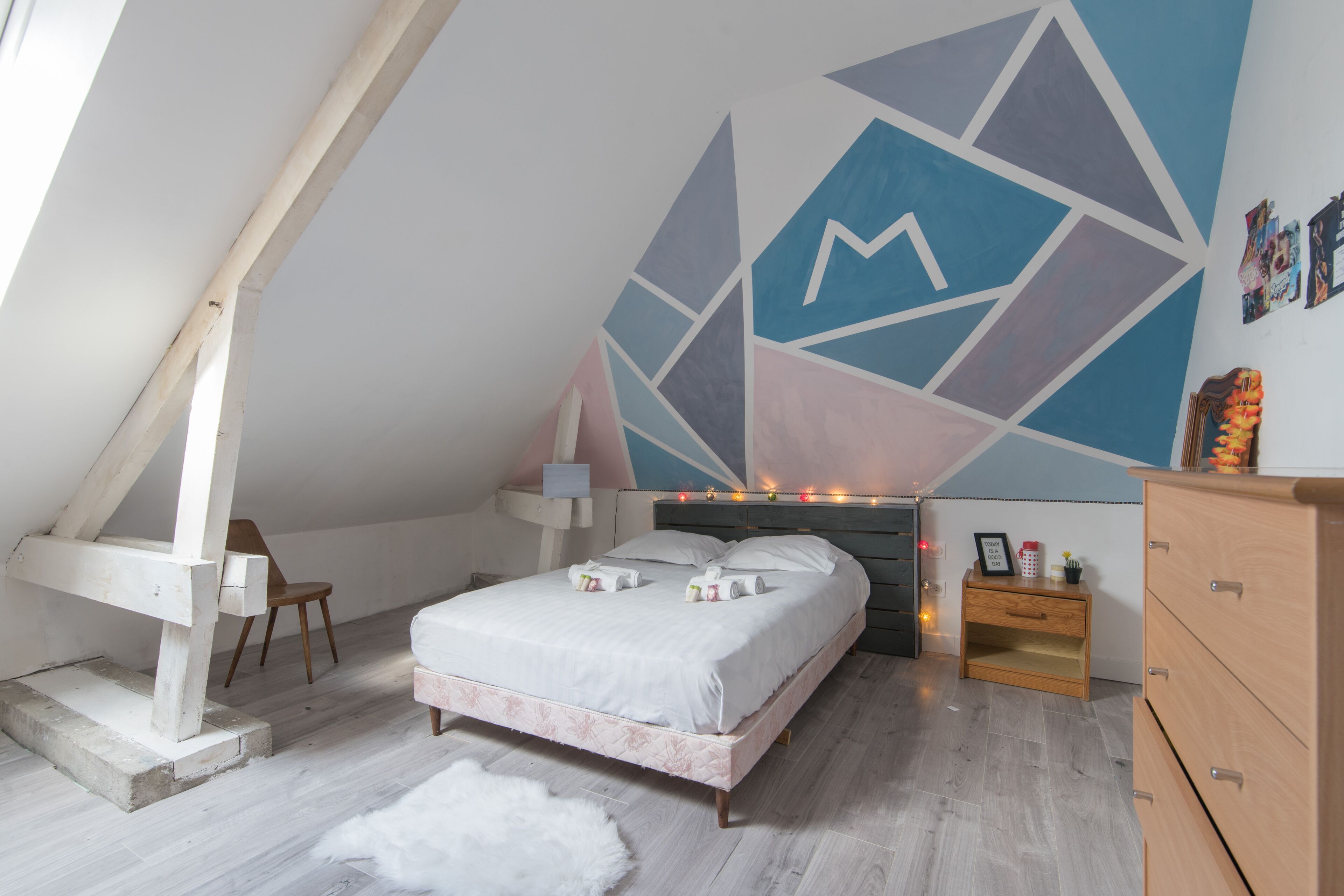 A bedroom with a sloped ceiling, a bed with white bedding, a wooden nightstand, and a geometric wall mural.