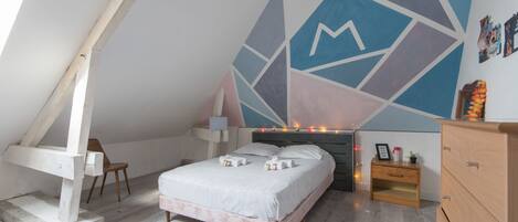 A bedroom with a sloped ceiling, a bed with white bedding, a wooden nightstand, and a geometric wall mural.