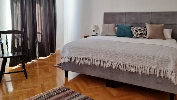 A bedroom with a bed, a chair, a rug, and a window with curtains. - Old bridge (Mostar)