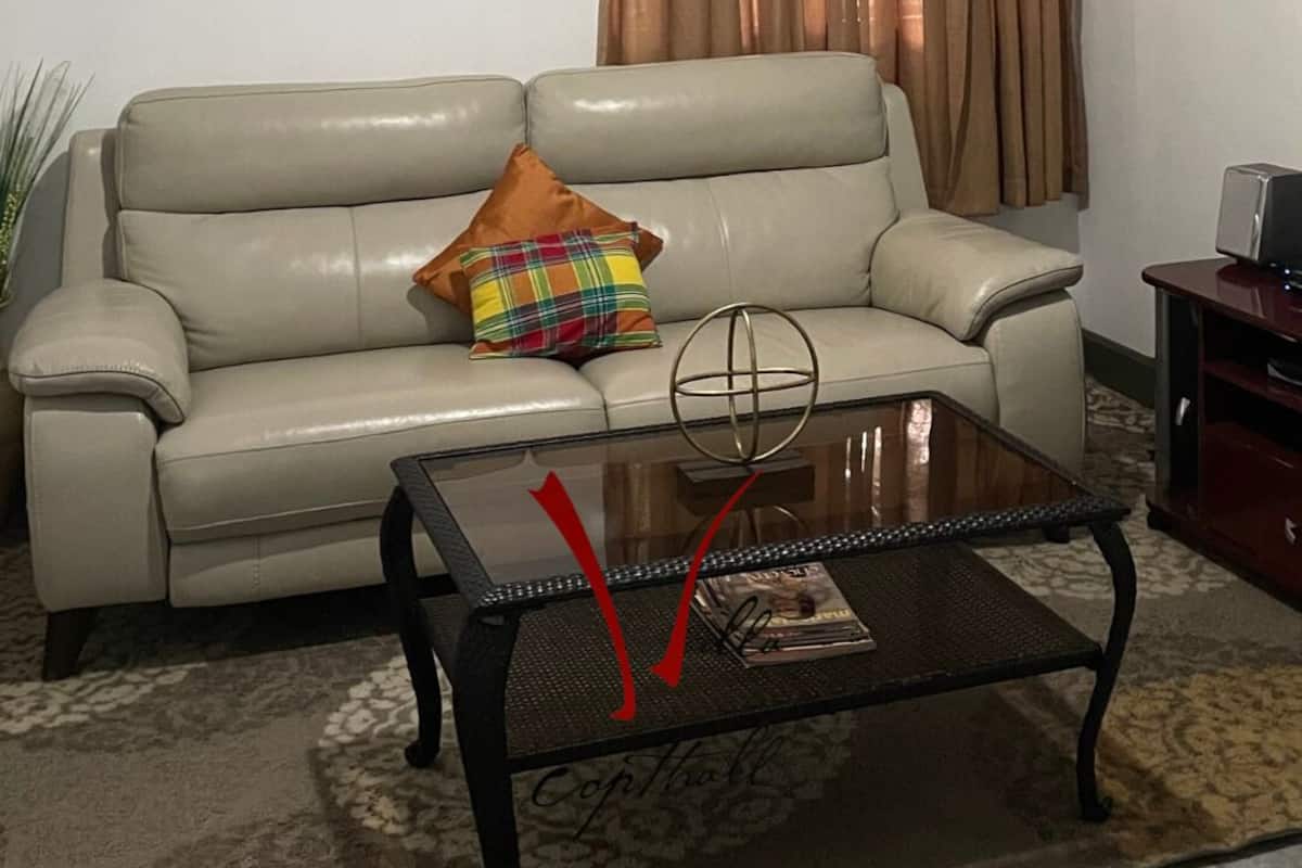 Sala de estar con un sofá color beige, una mesa de centro de vidrio, un ventilador de techo, una cortina marrón y un televisor montado en la pared.