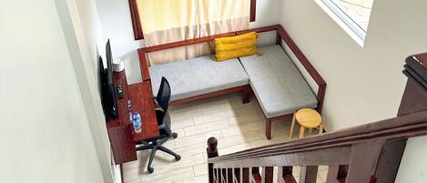 A compact room with a bed, a desk, a chair, a small stool, and a window.