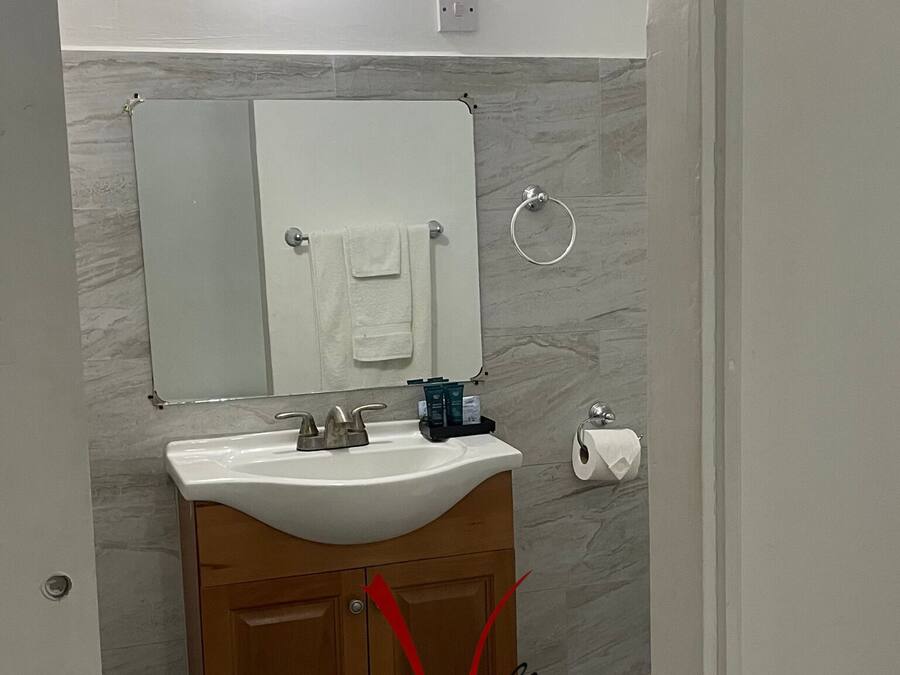 Un baño con lavamanos blanco, tocador de madera, un espejo, un aro para toallas y un sujetapapeles.
