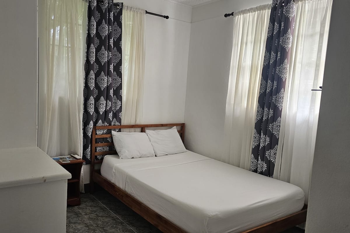 Una cama individual con ropa de cama blanca, cabecera de madera y dos almohadas blancas. Hay una mesita de noche con un control remoto y una botella de agua. La habitación tiene paredes blancas y una ventana con cortinas blancas traslúcidas y cortinas estampadas.