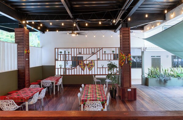 An outdoor dining area with wooden tables and chairs, string lights, and a view of a building in the background.