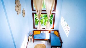 A small, bright room with a bed, a window with curtains, a small table, and a wall decoration.