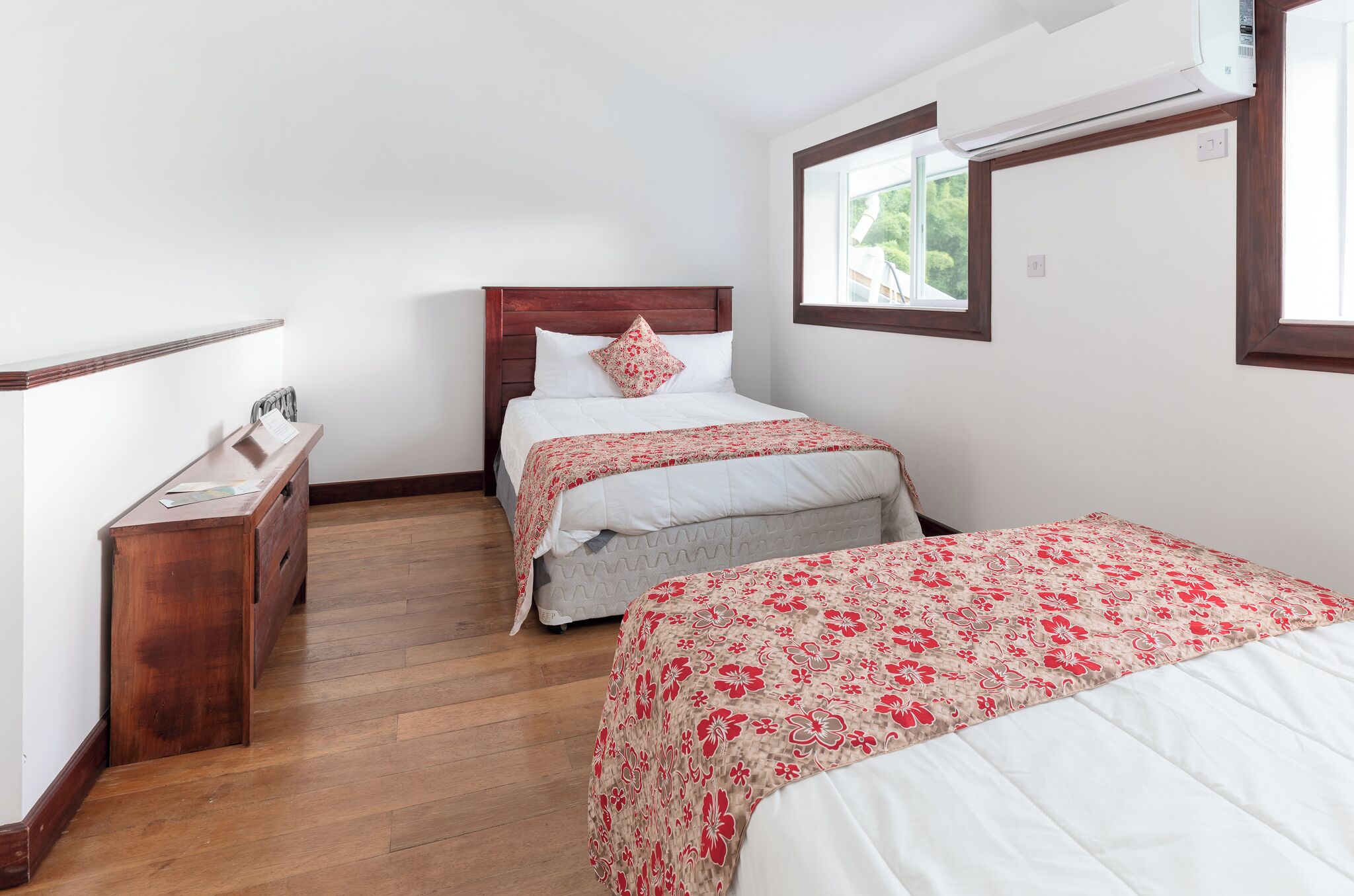 A bedroom with two beds, a wooden dresser, and a window with a view of greenery.