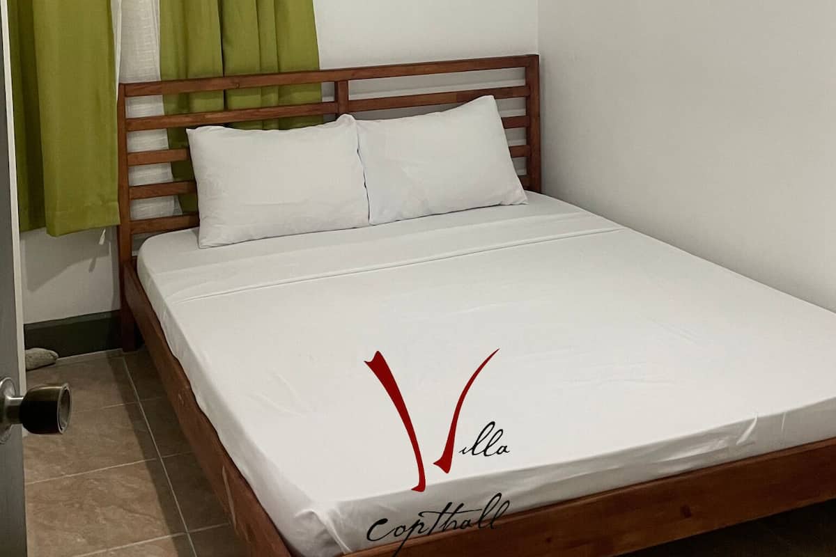 Una cama individual con estructura de madera y ropa de cama blanca en una habitación con cortina verde y paredes blancas.