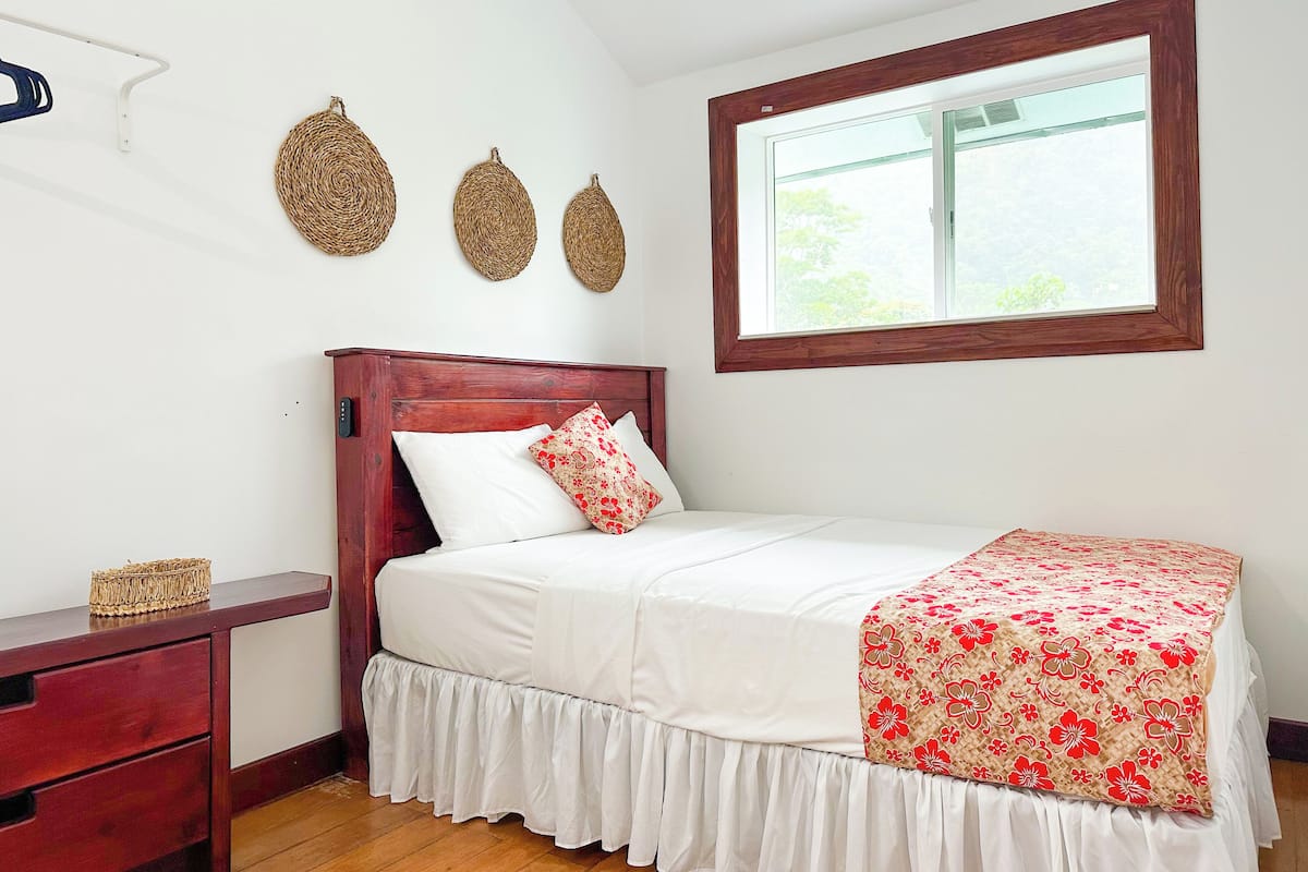 Un dormitorio ordenado con una cama de madera, ropa de cama blanca, una mesita de noche con una cesta y una ventana con vistas a zonas verdes.