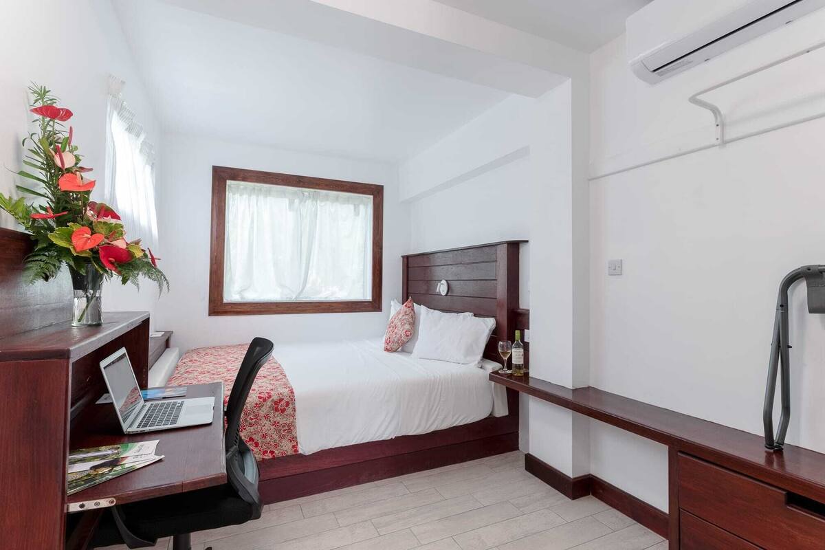 Habitación de hotel con cama, escritorio con ordenador portátil, jarrón con flores y ventana con cortinas.