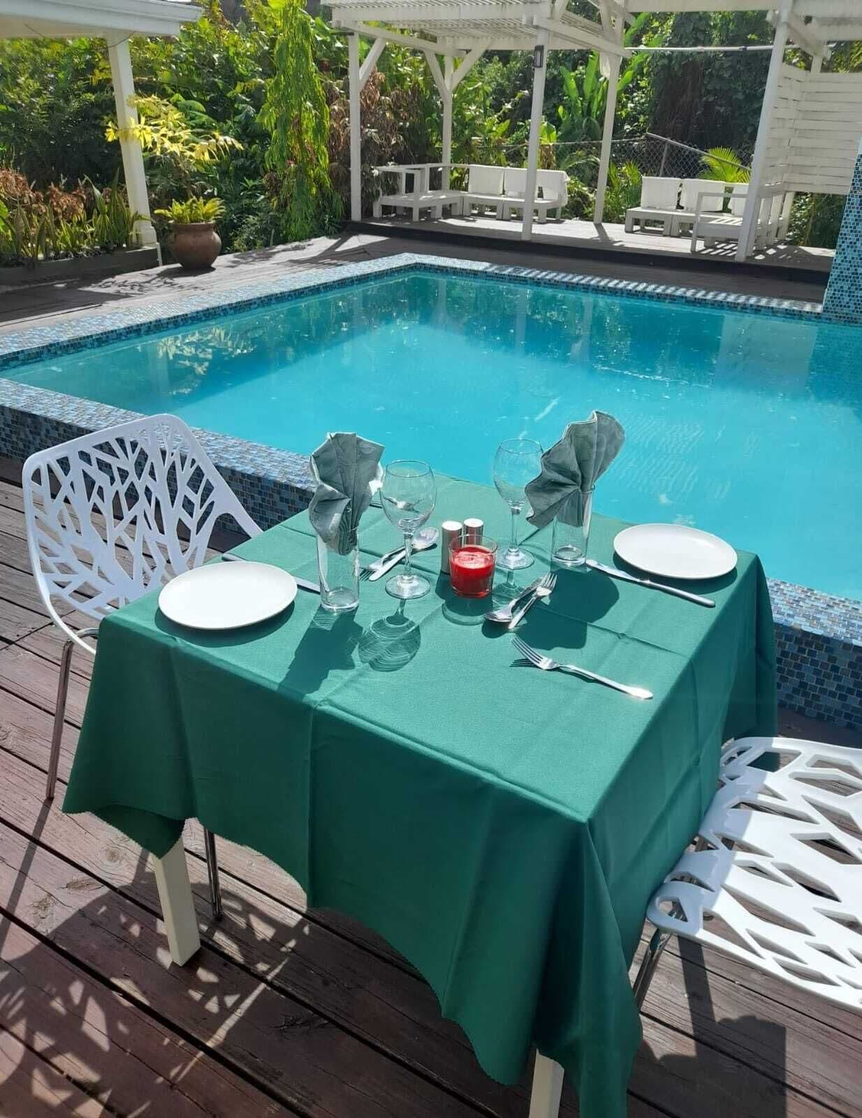 En spiseplads ved poolen med et bord til to, omgivet af en hvid pergola og frodigt grøntværk.