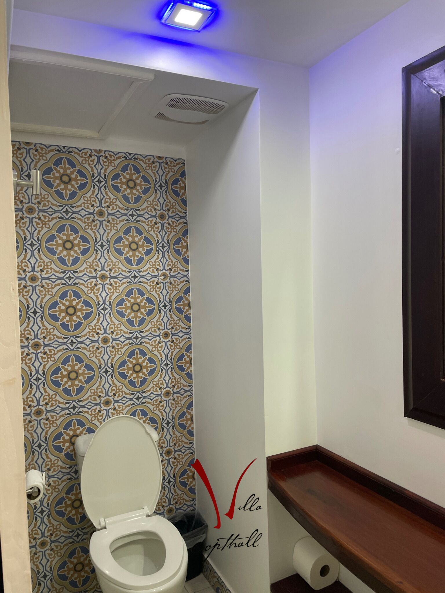 Un bagno con parete rivestita di piastrelle decorate, un WC bianco, uno scaffale in legno e un cartello con la scritta "Villa Opti hall".