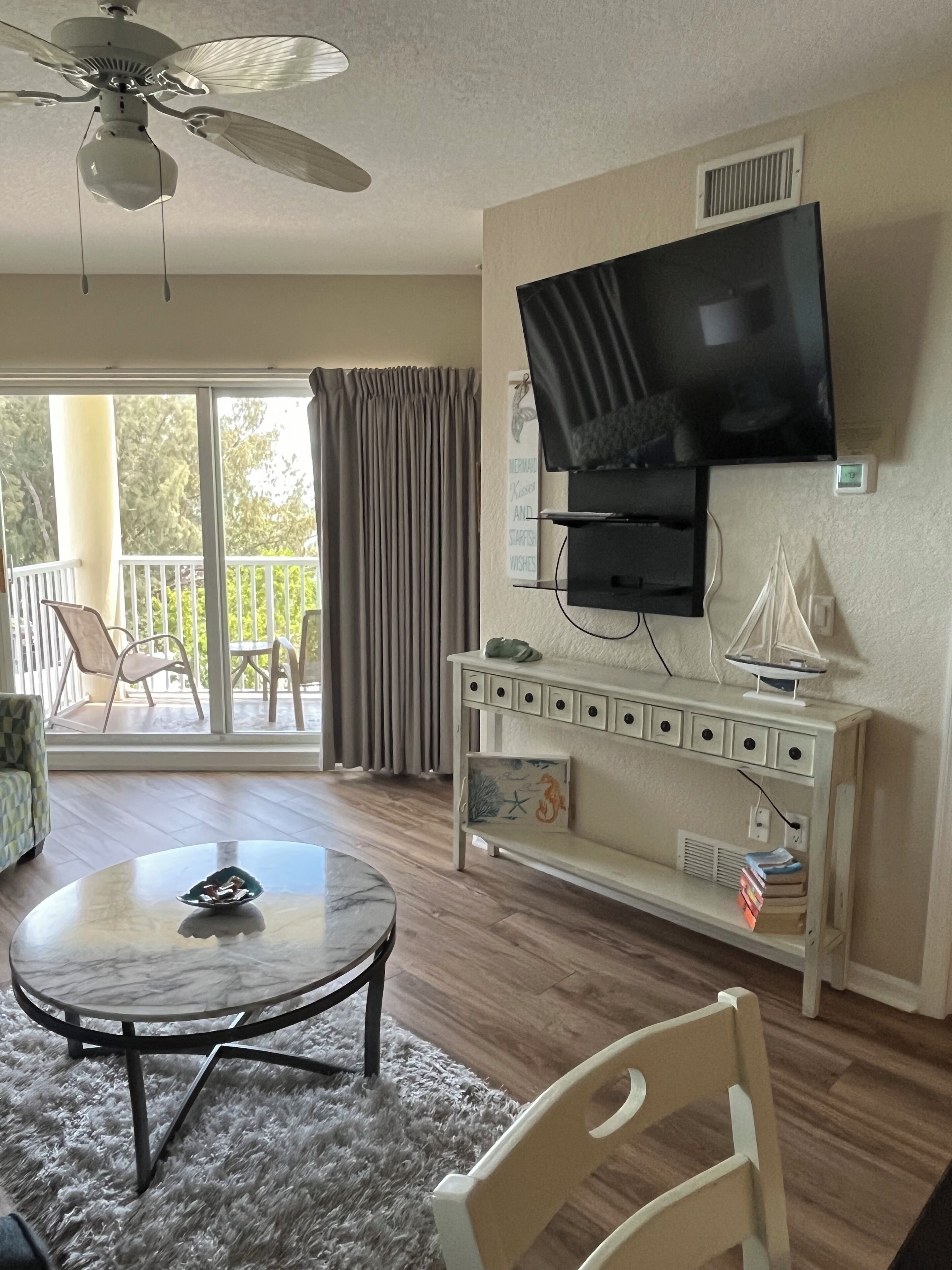 A living room with a round table, a television mounted on the wall, a ceiling fan, and a view of the outdoors through large windows.
