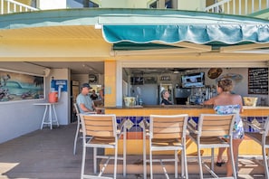 An outdoor bar area with wooden chairs, a counter, and a television screen. - Condo on the beach! 2/2 with two pools, cafe and tiki bar! Sunset Vista Resort (Treasure Island)