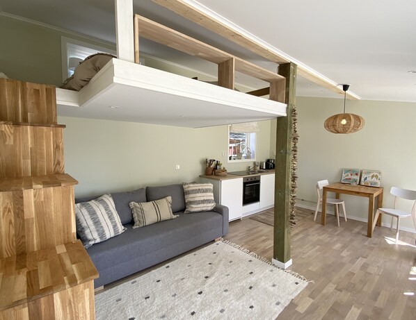 A modern living room with a grey sofa, wooden staircase, and a kitchen area with white cabinets and a dining table.