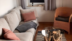 A living room with a beige sofa, a wicker chair, a glass coffee table, and a window with curtains. - Aalhaus Wustrow apartment to the meadow 900 m to the beach (Wustrow)