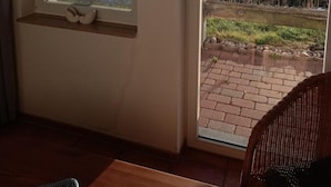 A room with a large window overlooking a grassy field and a body of water. There is a wicker chair and a small table in the foreground. - Aalhaus Wustrow apartment to the meadow 900 m to the beach (Wustrow)