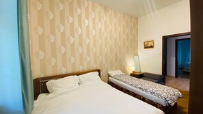 Habitación de hotel con una cama grande, una cama más pequeña, una mesita de noche con lámpara, un cuadro en la pared y una puerta que da a otra habitación.