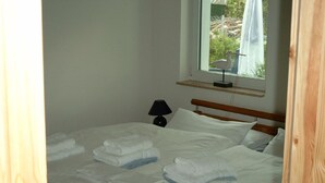 Una cama bien hecha con sábanas y toallas blancas, una lámpara de noche y una ventana con vistas a follaje.