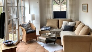 Una sala de estar con sofá, sillón y reposapiés, una mesita de madera y una ventana con cortinas.