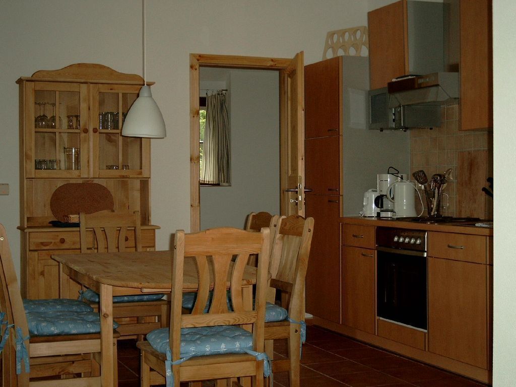 A wooden dining table with chairs, a kitchen with wooden cabinets, a stove, and a hanging light fixture.