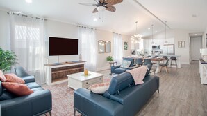 A modern living room with a large TV, blue sofas, a coffee table, and a ceiling fan. - Brand new home, 1 block from Beach (Gulfport)